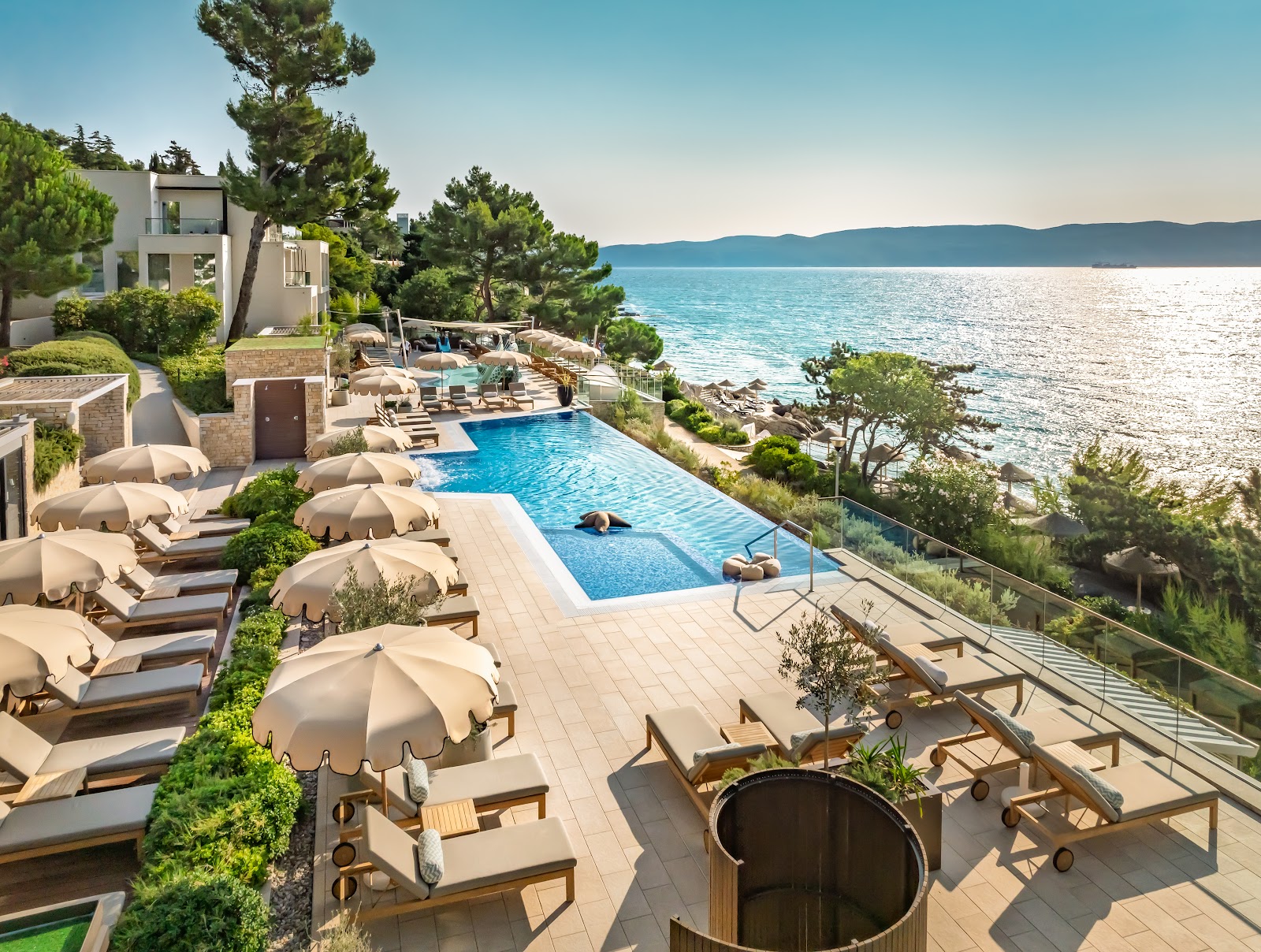 Pool deck with beige umbrellas and lounge chairs overlooking the Adriatic Sea and distant mountains at Girandella Resort.