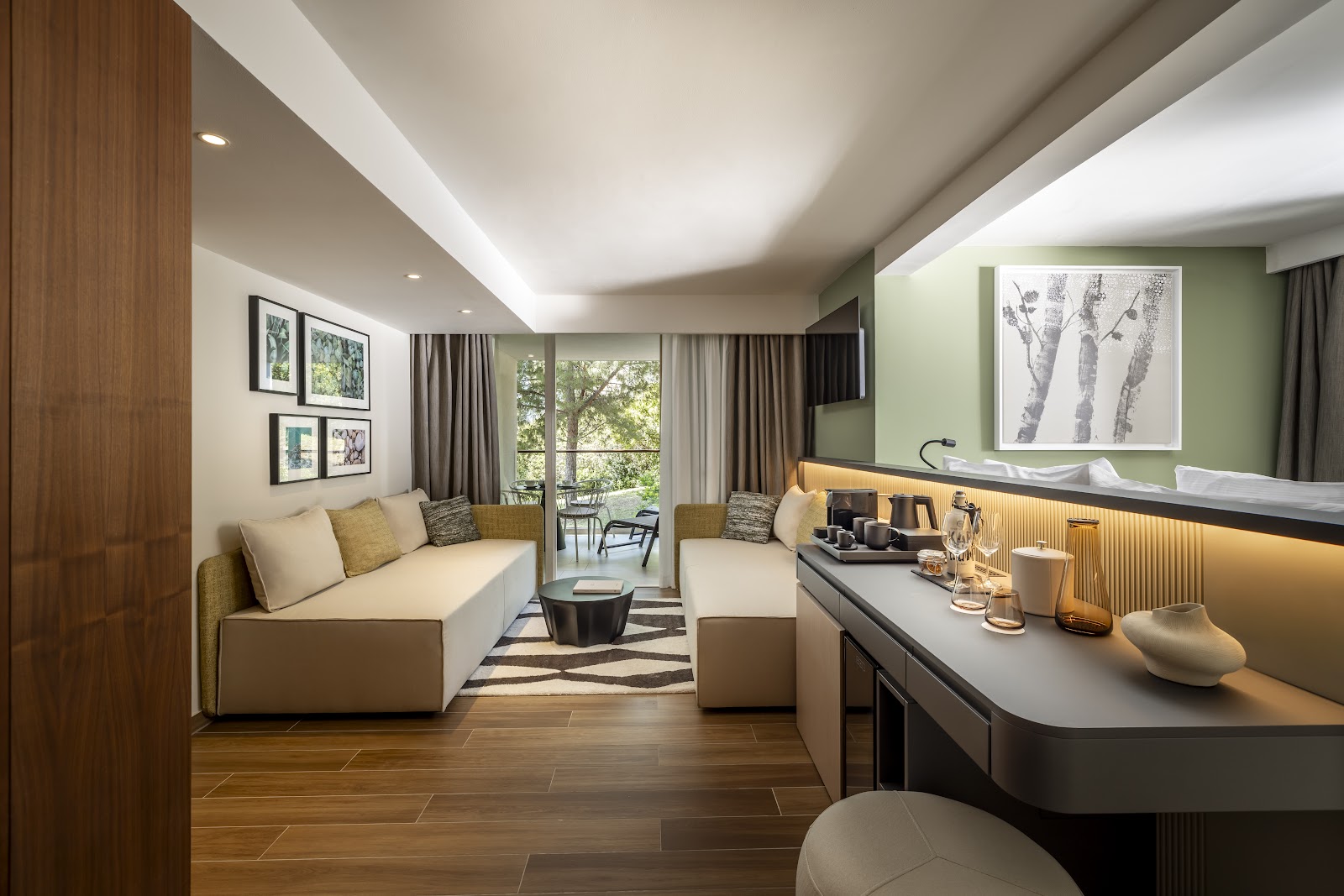 Modern resort suite with wood flooring, seating area with cream sofa, green accent wall, vanity counter, and balcony overlooking trees.