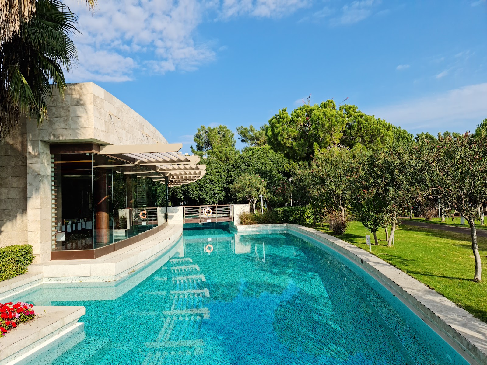 Rectangular turquoise pool lined with lounge chairs, bordered by a modern stone pavilion with pergola and manicured lawn with mature trees under blue sky.