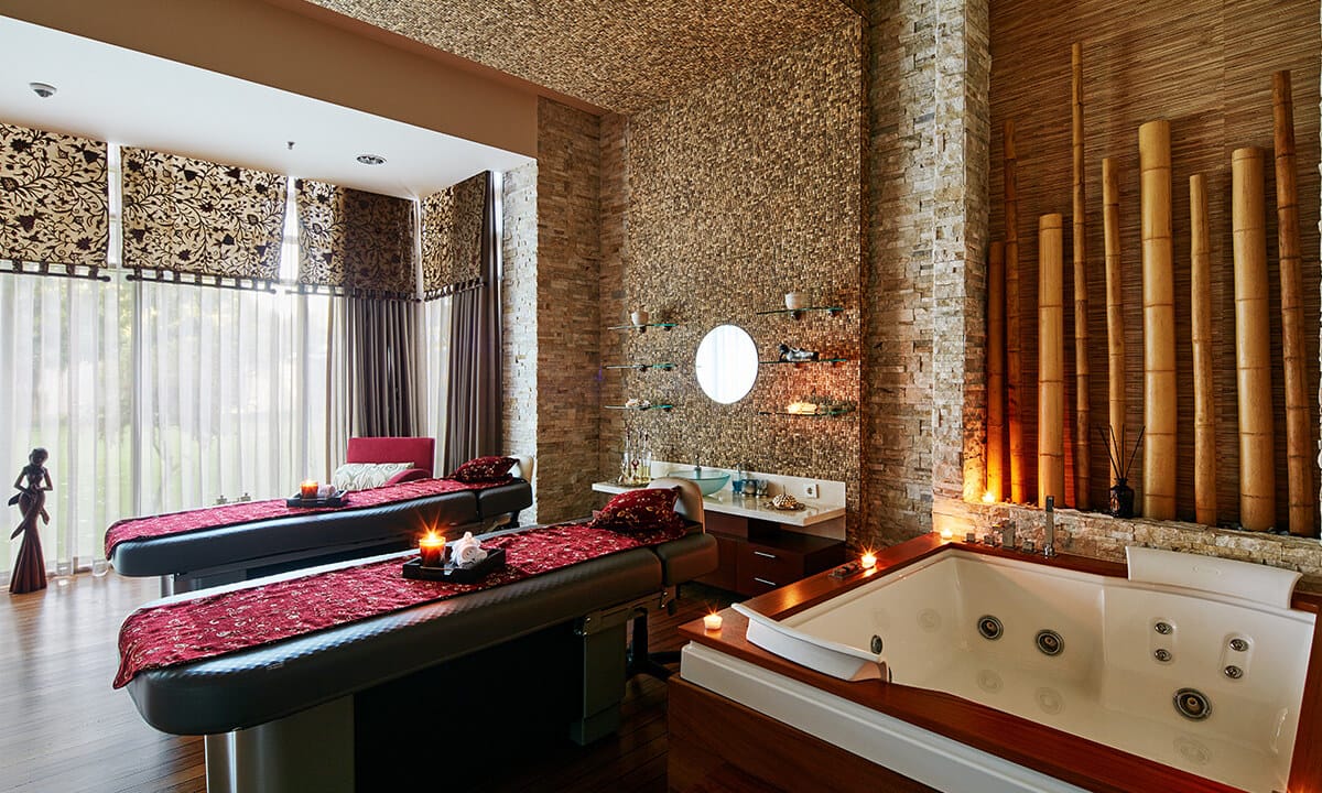 Spa treatment room with two massage tables, a sunken whirlpool tub, brick walls, tall bamboo accents, and candlelit ambiance.