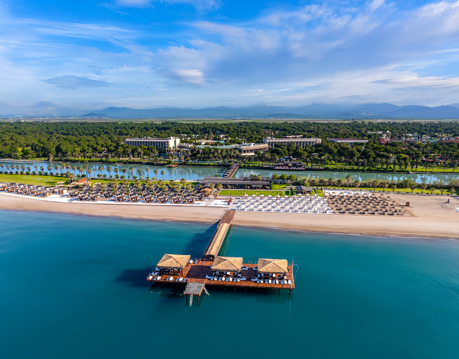 Aerial view of Gloria Serenity Resort's beachfront with wooden pier, turquoise sea, sandy beach with loungers, and riverside lagoon beyond.