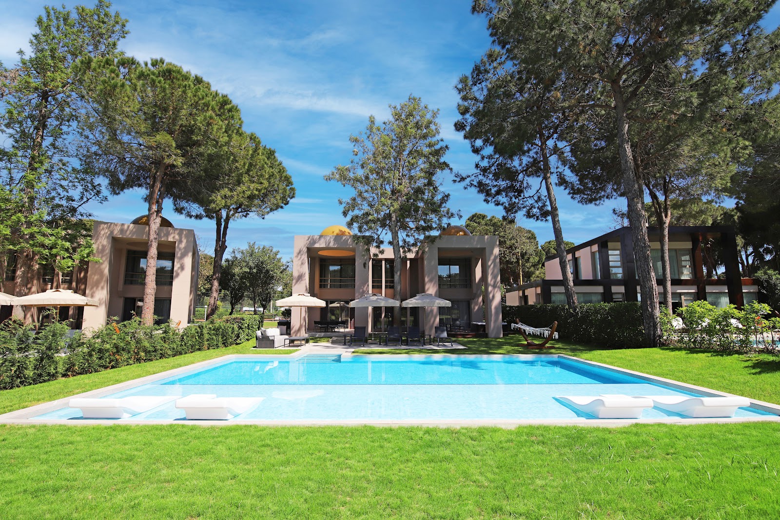Modern stone resort buildings with blue rectangular pool, loungers, and mature pine trees on manicured lawn under clear sky.
