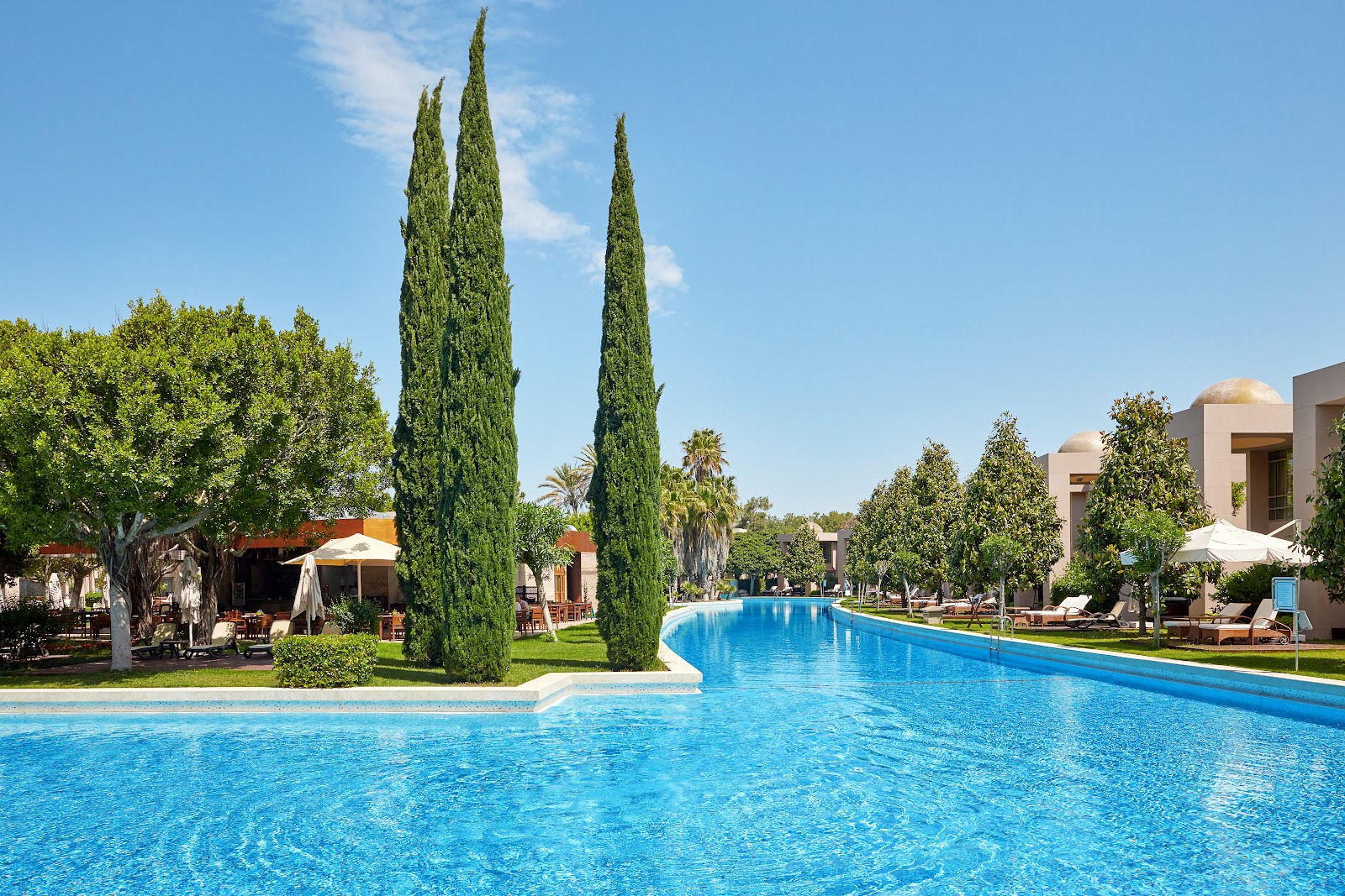 Tall cypress trees frame a clear blue pool lined with loungers and umbrellas at Gloria Serenity Resort, with whitewashed buildings and manicured gardens beyond.