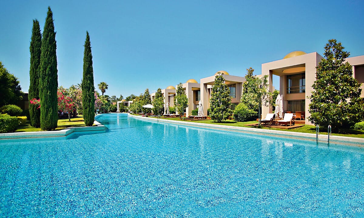 Long rectangular pool with turquoise water flanked by beige villa suites, cypress trees, and manicured lawns under clear blue sky.