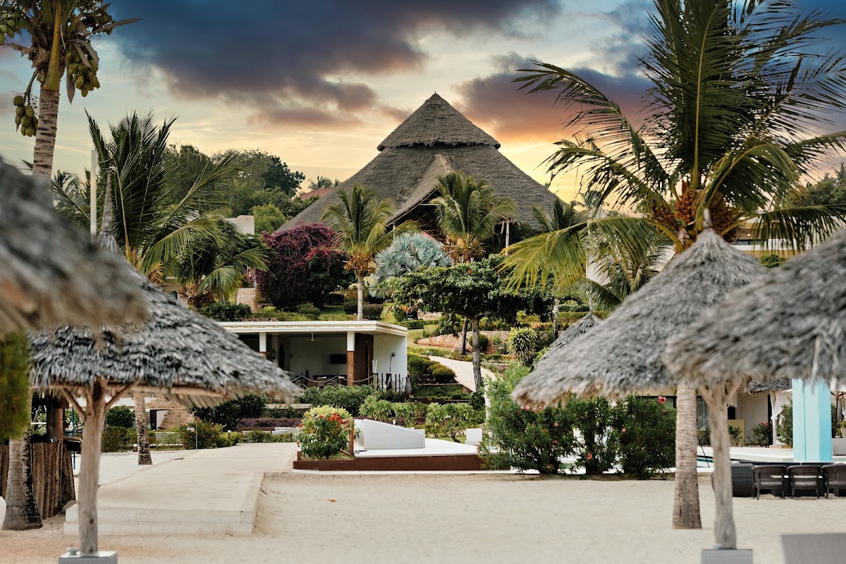 Sandy beach area with thatched-roof bungalows and a large central structure with conical roof, surrounded by palm trees at sunset.