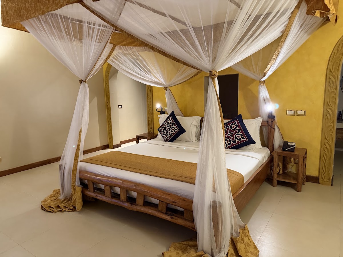 Four-poster canopy bed with white drapes and decorative navy pillows in a yellow room with wooden furniture.