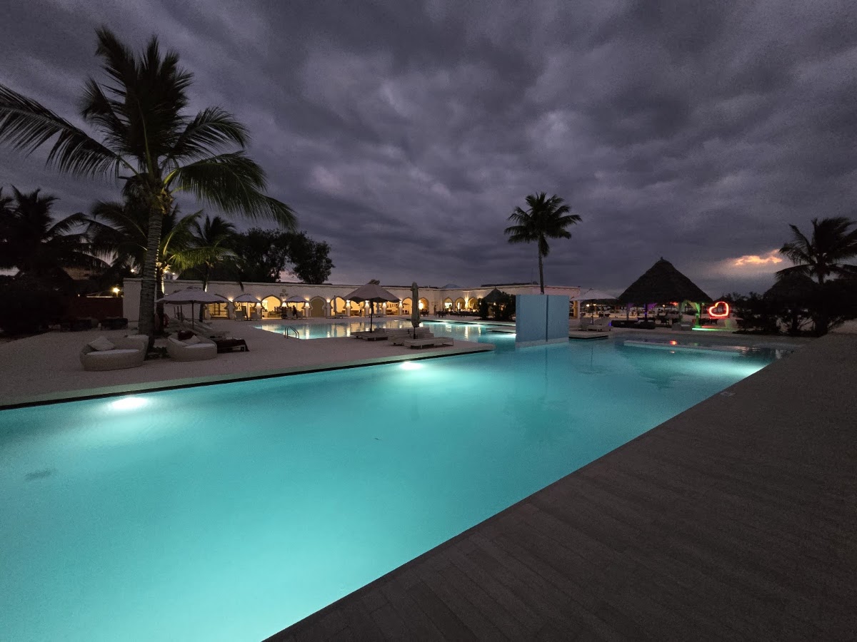 Illuminated turquoise pool at dusk with lit dining pavilion, palm trees, and thatched-roof structures under stormy sky at Gold Zanzibar Beach House & Spa.