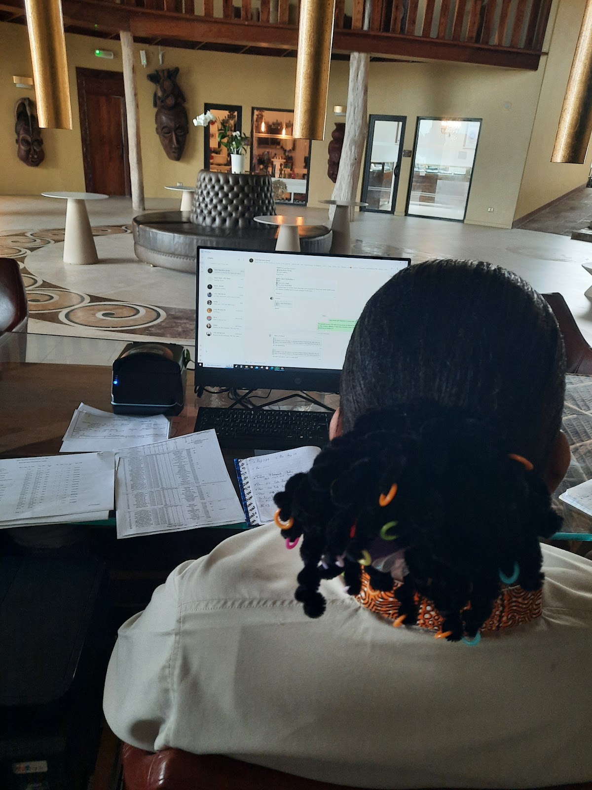 Staff member works at a desk in the Gold Zanzibar Beach House & Spa lobby, with tall columns, African masks, and decorative elements visible in the background.