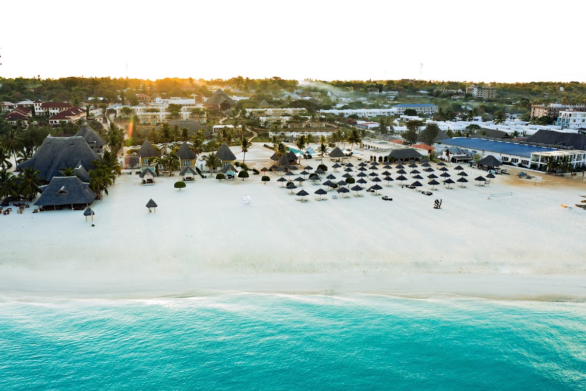 Aerial view of Gold Zanzibar Beach House & Spa with turquoise waters, white sand beach lined with black umbrellas, and thatched-roof buildings among residential homes.