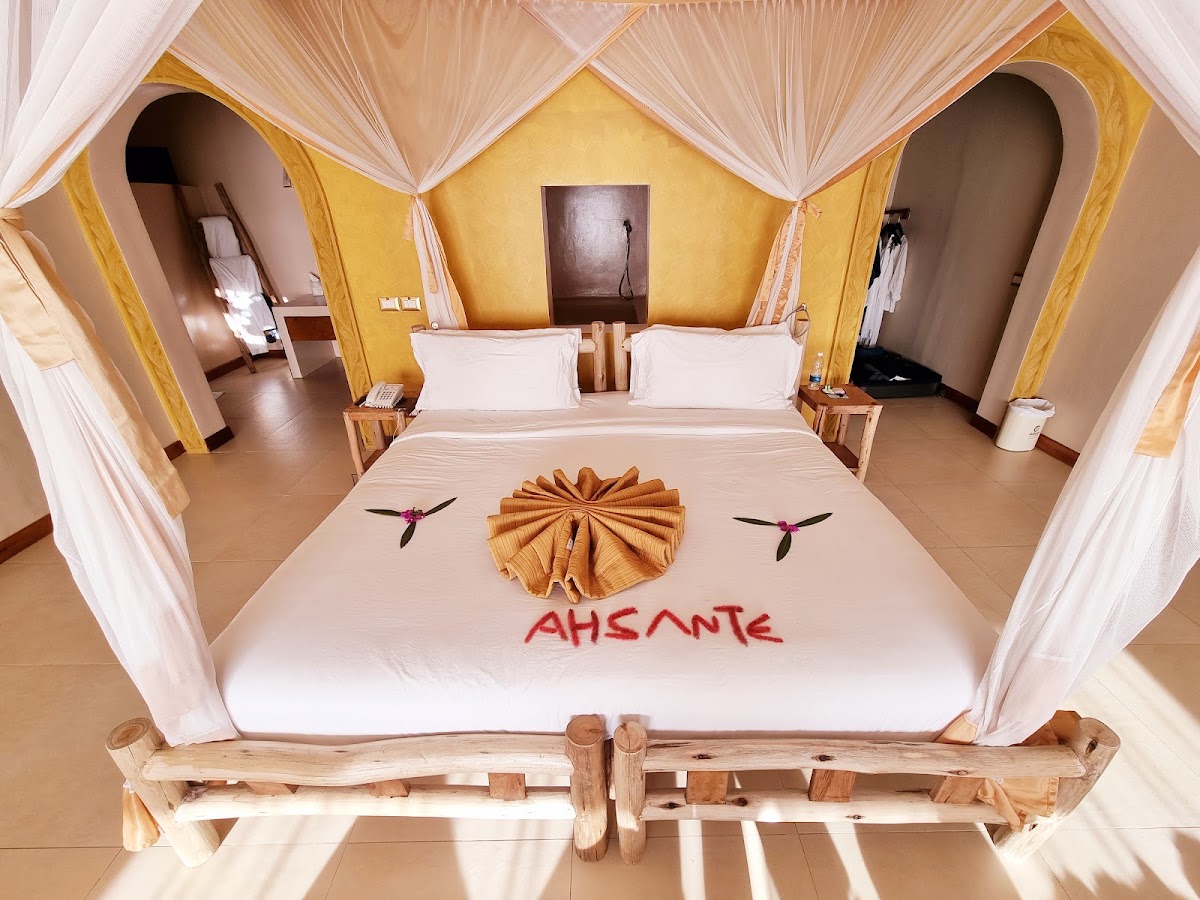 Bedroom at Gold Zanzibar Beach House & Spa with yellow walls, white sheer canopy drapes, and a bed decorated with a shell-shaped towel arrangement and text.