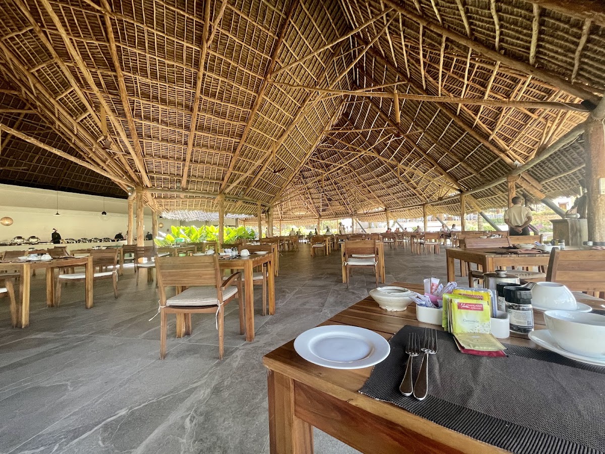 Open-air dining pavilion with woven palm-thatch roof, wooden tables and chairs, place settings with white plates, and staff in the background at Gold Zanzibar Beach House & Spa.