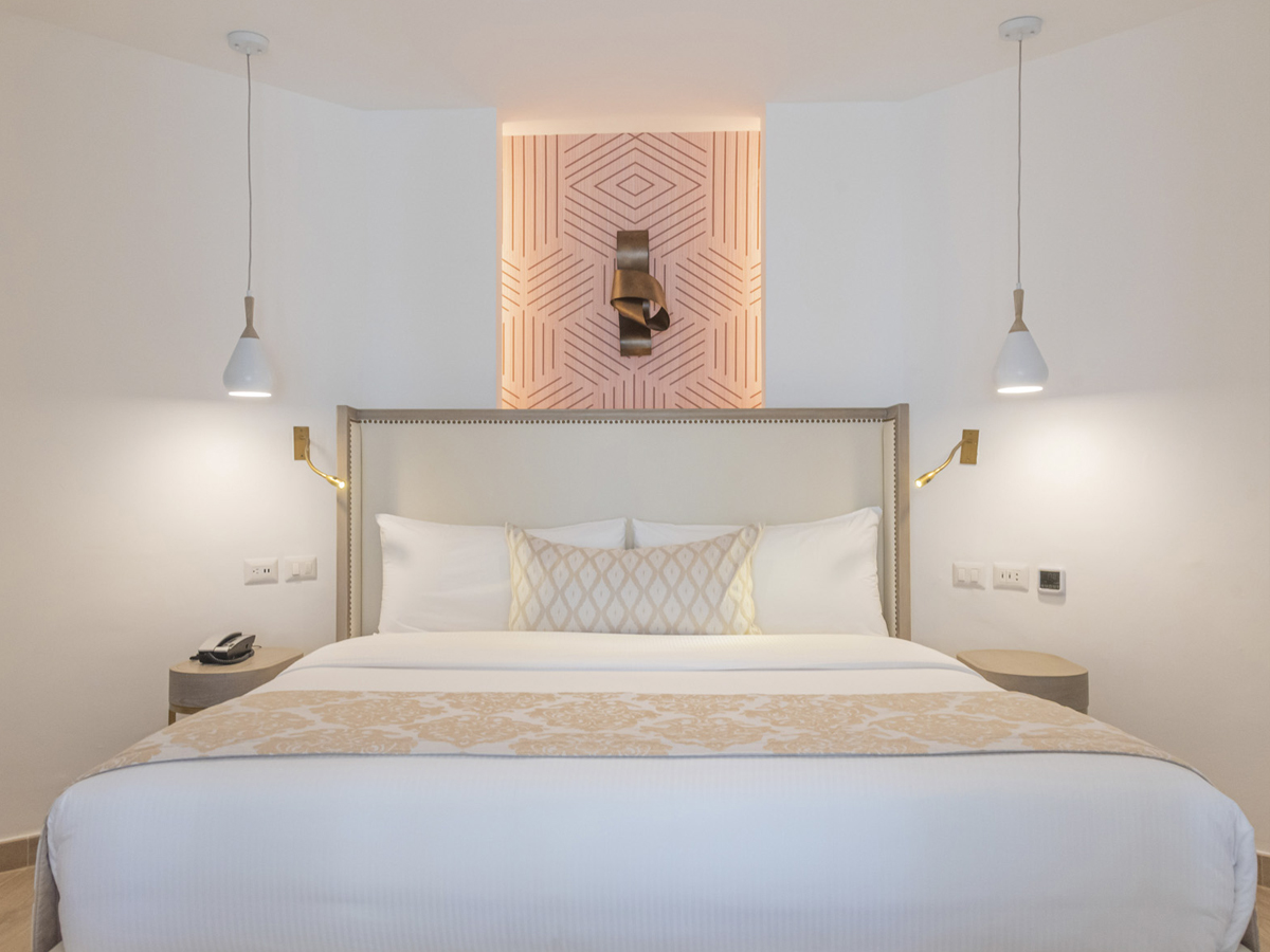 Modern resort bedroom with king bed, upholstered headboard, pendant lights, and geometric wall art above at Grand Bávaro Princess.