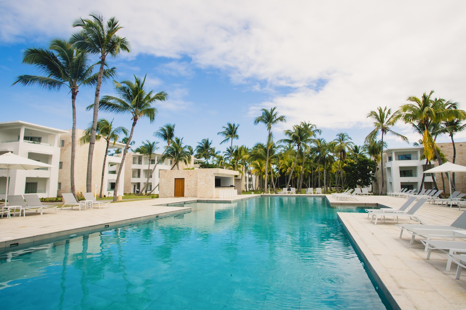 Large rectangular pool with turquoise water surrounded by white lounge chairs, palm trees, and modern white buildings at Grand Bávaro Princess.