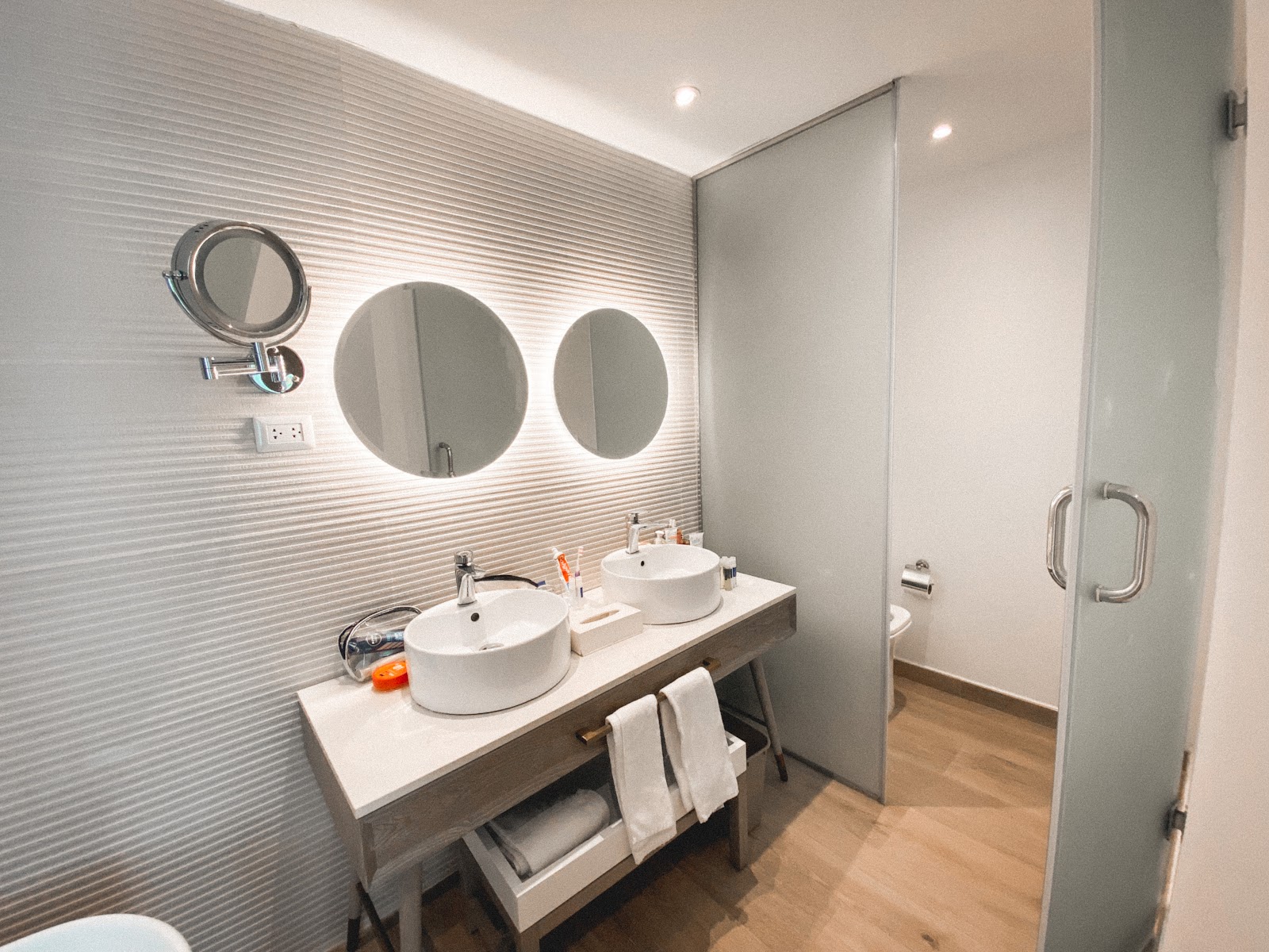 Bathroom with two round illuminated mirrors above white vessel sinks on a gray vanity, textured white wall tiles, and wood flooring.