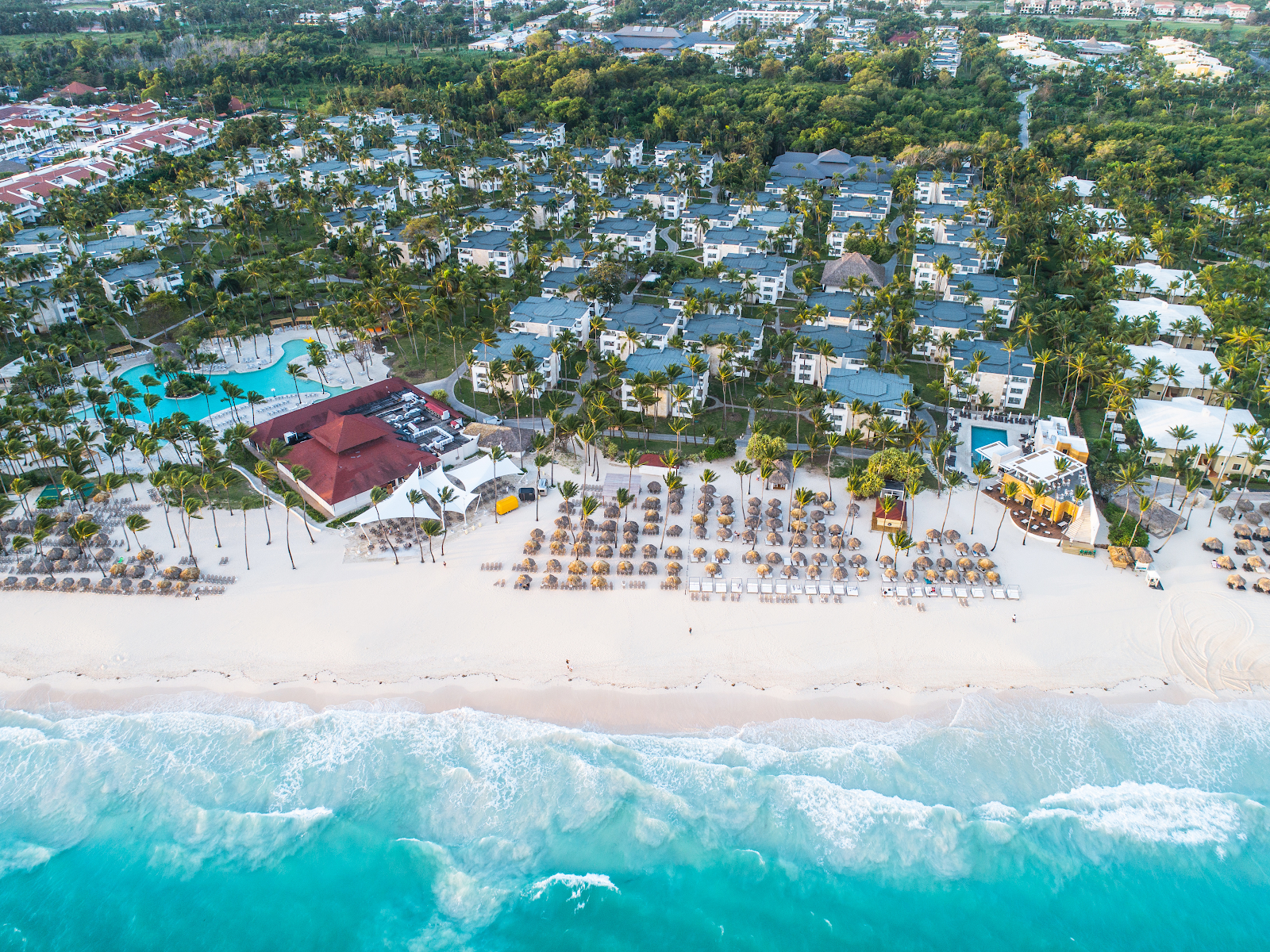 Aerial view of Grand Bávaro Princess with turquoise ocean waves, white sand beach lined with lounge chairs, red-roofed main building, palm trees, and white resort buildings scattered inland.