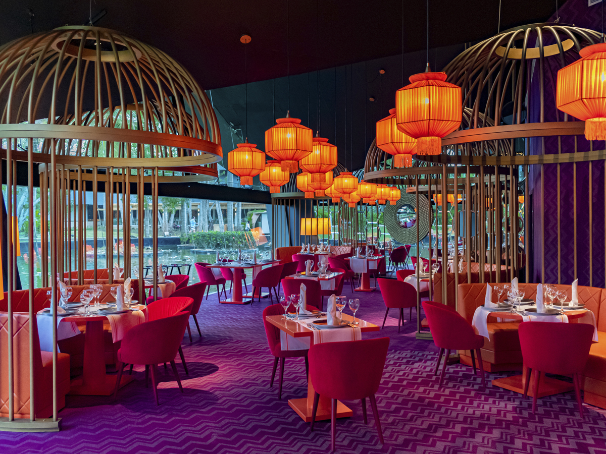 Modern dining room with red velvet chairs, wooden lattice partitions, hanging orange lanterns, and floor-to-ceiling windows overlooking tropical grounds at Grand Bávaro Princess.