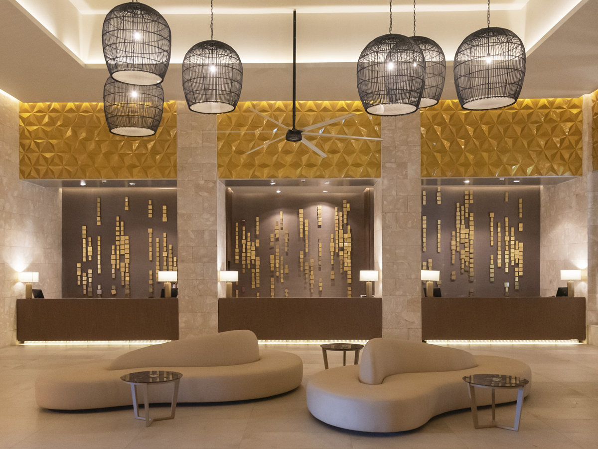 Modern resort lobby with curved seating, hanging woven pendant lights, and three reception desks with gold tile accents behind them.