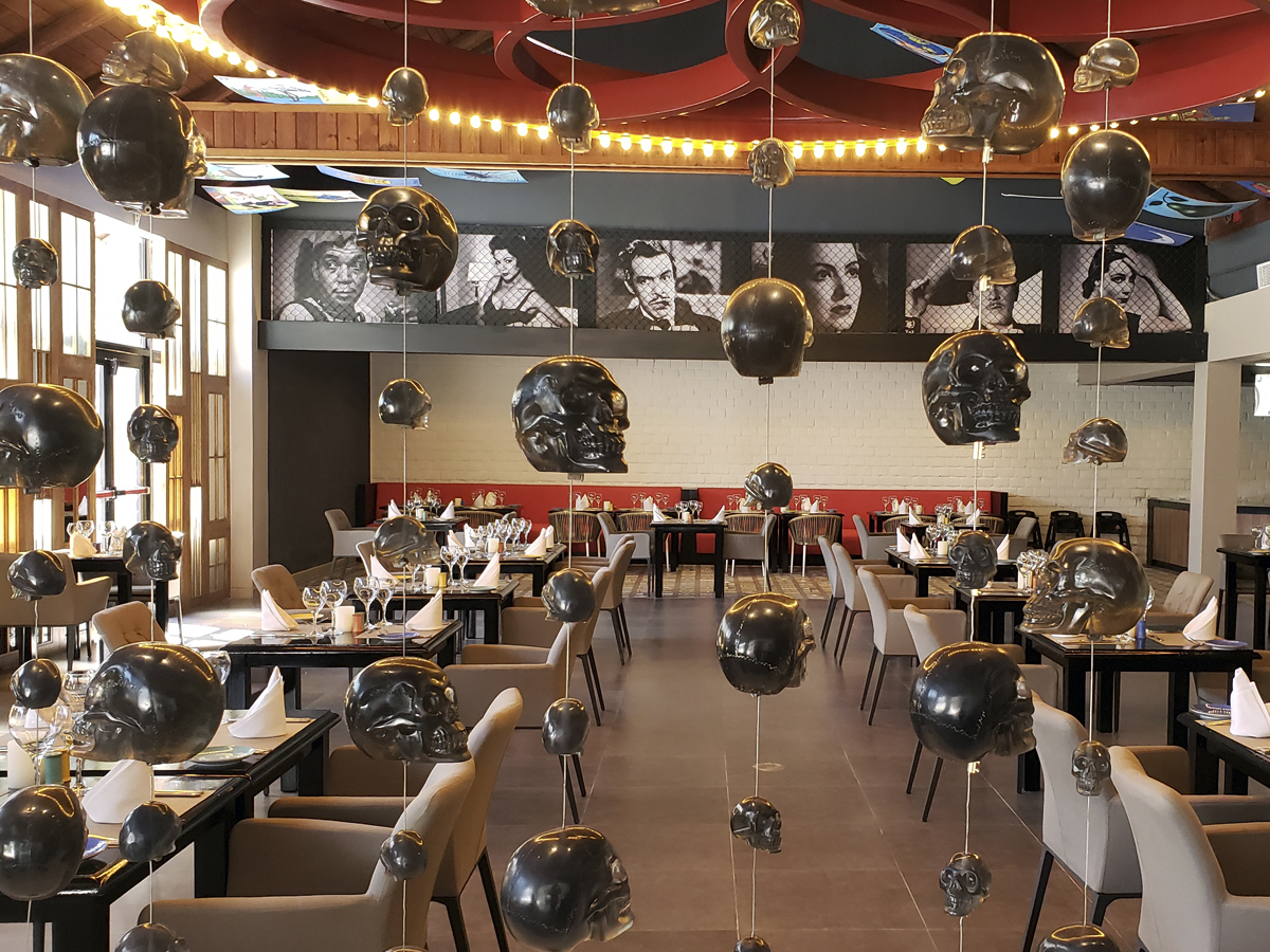 Dining room with black tables, beige chairs, and hanging bronze skull sculptures under warm string lights, with black and white portrait photos on the back wall.