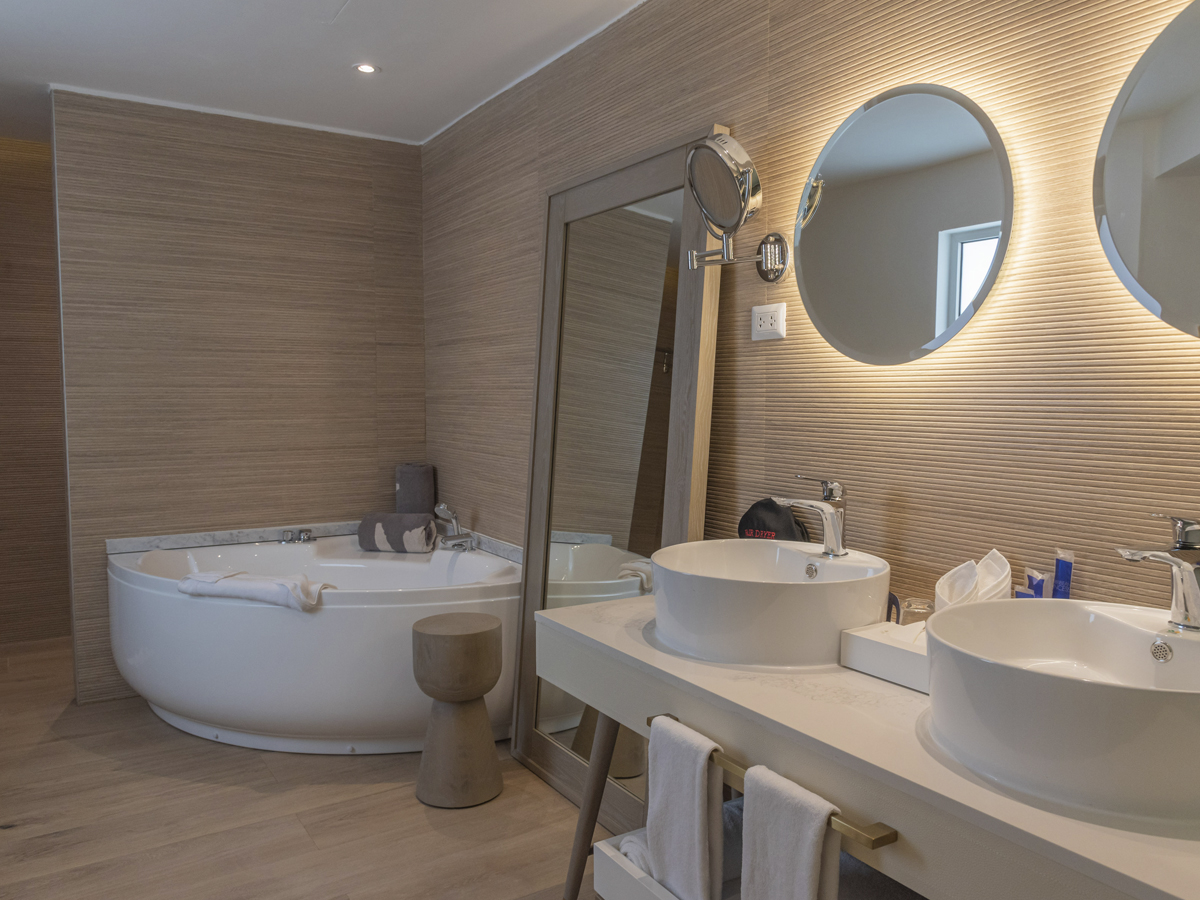 Spa-style bathroom with dual round vessel sinks, illuminated mirrors, soaking tub, and textured beige tile walls.
