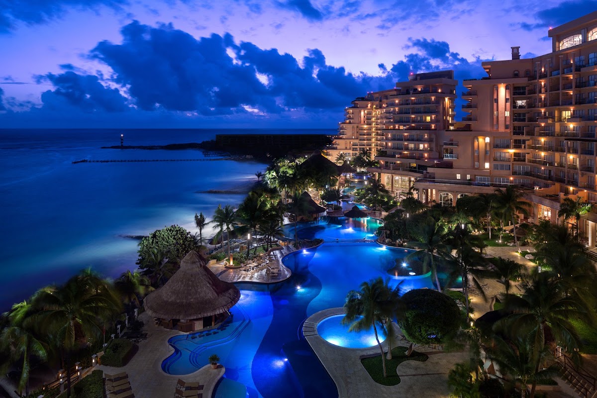 Grand Fiesta Americana Coral Beach Cancún at twilight, with illuminated pools, beachfront buildings, and dark ocean under purple-blue sky.