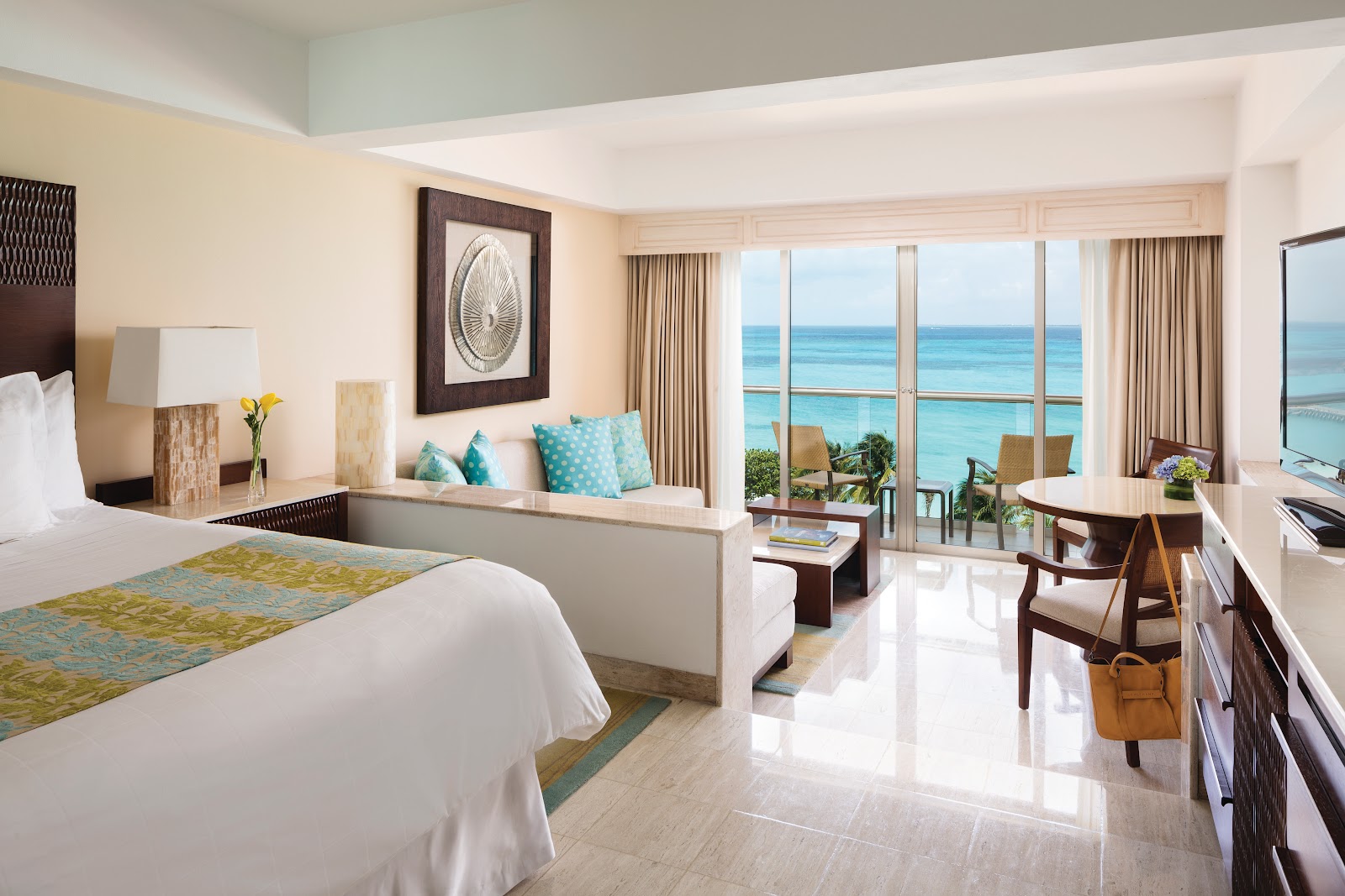 Modern resort bedroom with ocean view from balcony, white bed, turquoise accents, and tropical sea visible through large windows.