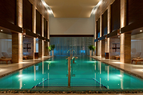 Indoor pool with blue-lit water and metal railings, flanked by tan lounge chairs and dark wood architectural features with ambient lighting.