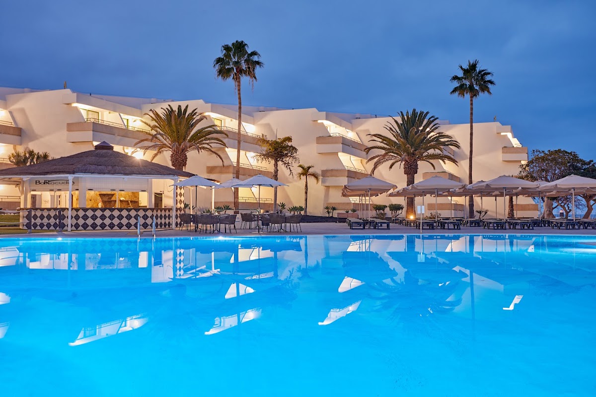 Illuminated resort buildings with stepped architecture overlook a bright blue pool lined with white umbrellas and palm trees at dusk.