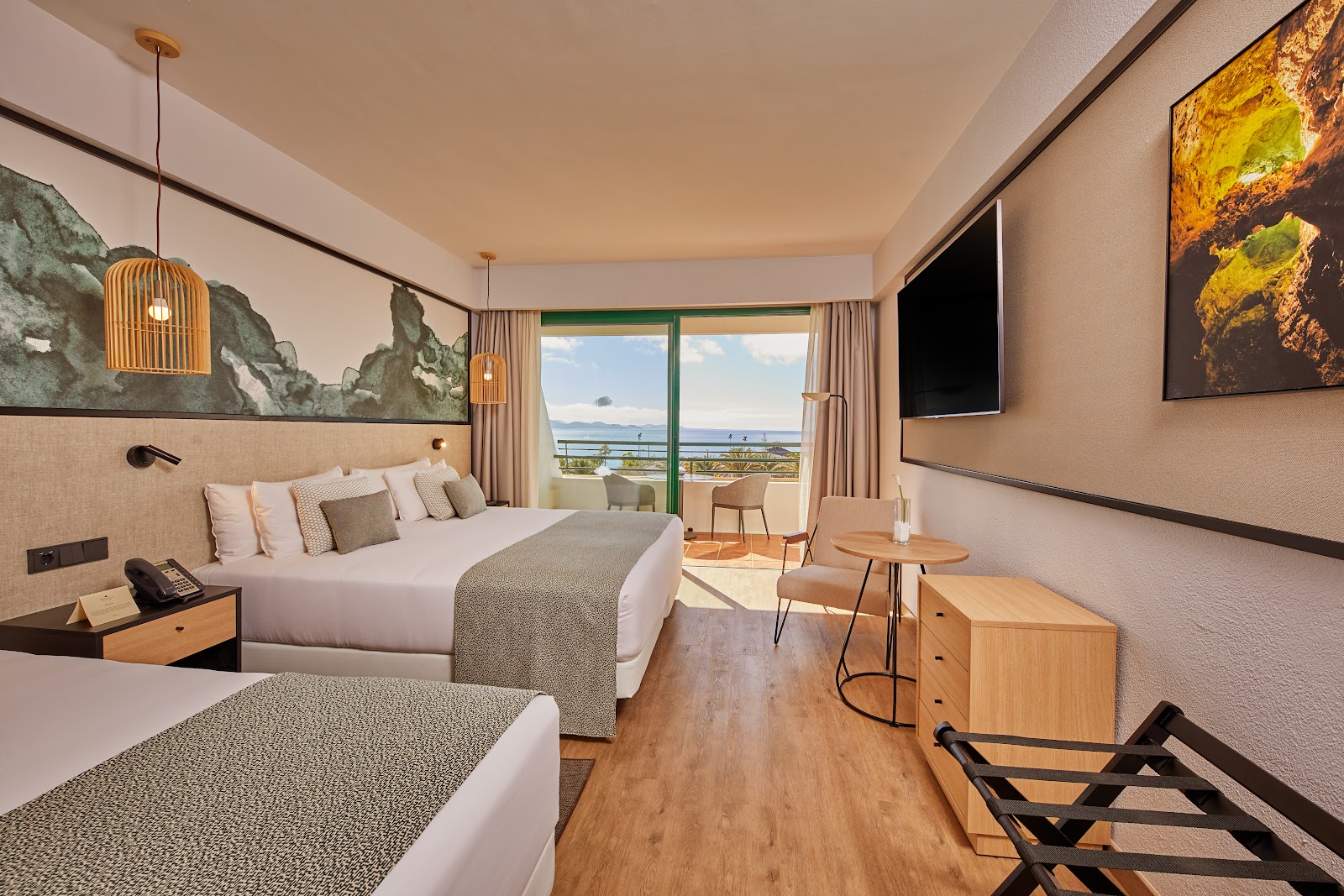 Modern hotel room with two beds, wooden furnishings, and a balcony overlooking the ocean and coastline.