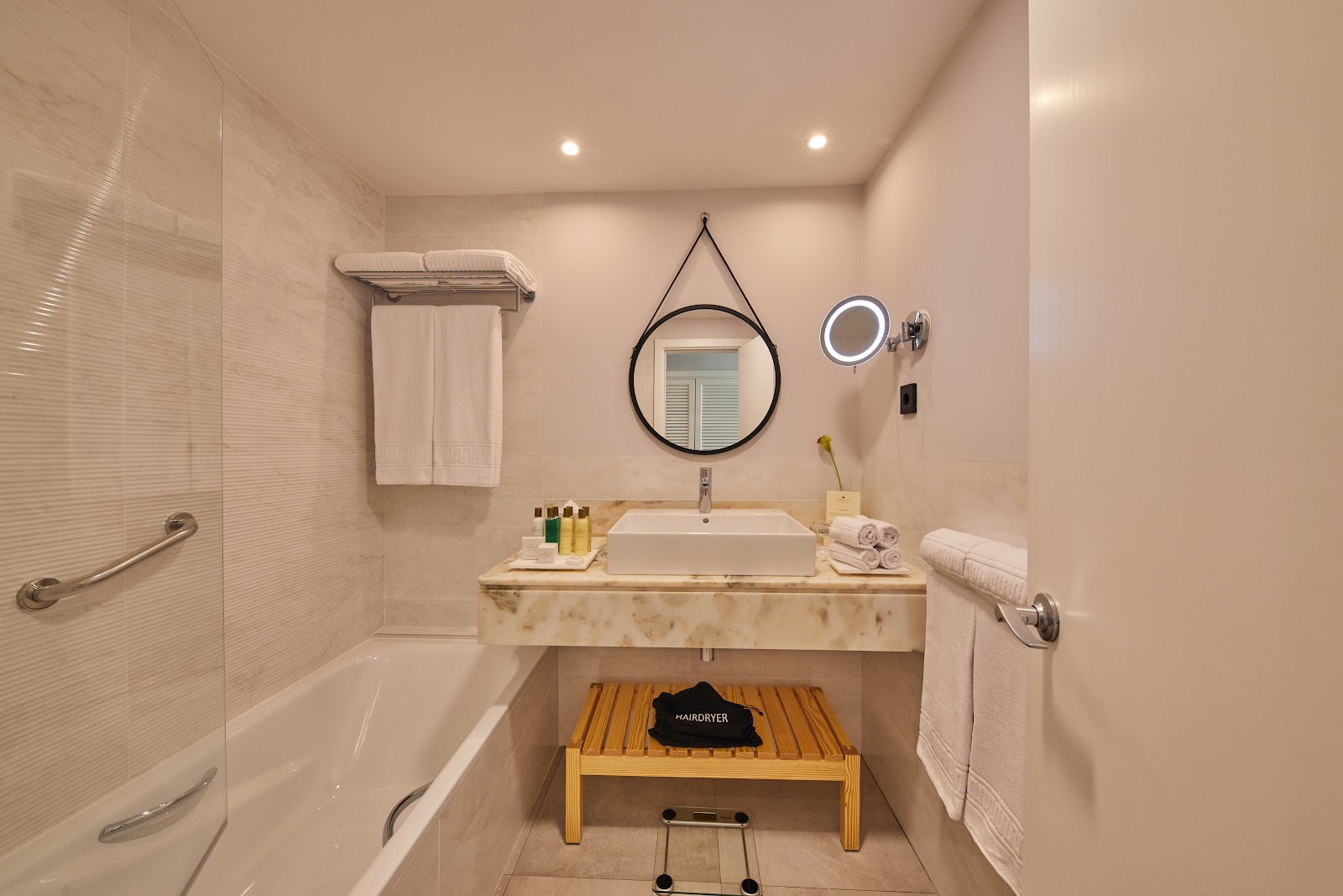 Modern resort bathroom with marble vanity, round hanging mirror, bathtub, rainfall shower, and wooden bench with hairdryer.