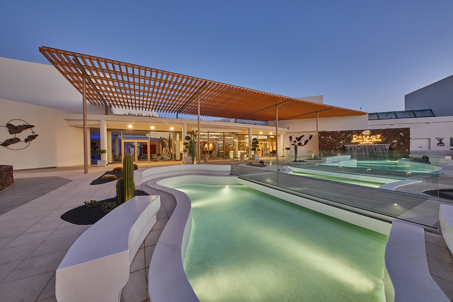 Illuminated thermal pools with curved white edges and green-tinted water sit on a terrace beside a modern building with wooden pergola at dusk.
