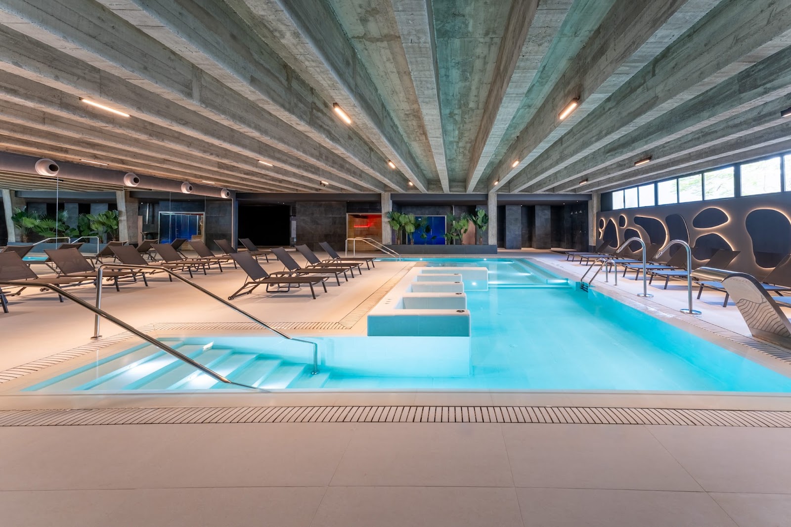 Indoor pool with turquoise water and tiered sun loungers on either side beneath a concrete ceiling with skylights at Grand Hyatt Lanzarote Playa Dorada Resort.
