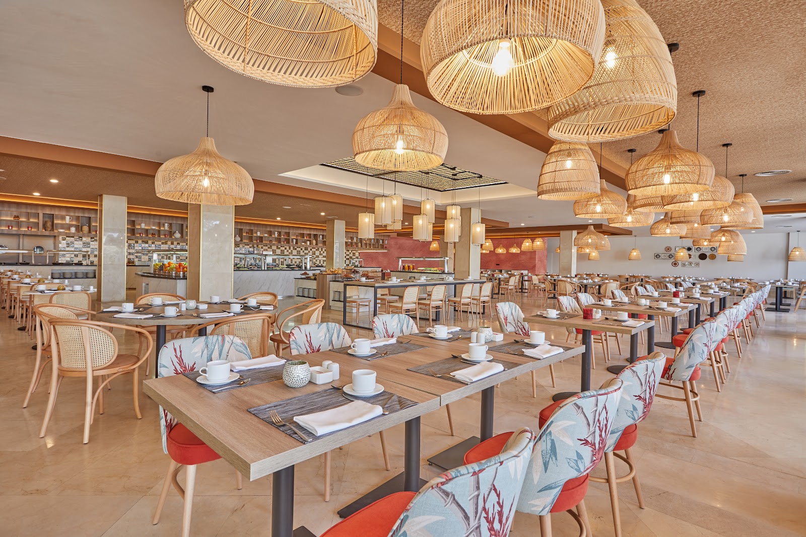 Spacious dining hall with woven pendant lights, wooden tables set with place settings, upholstered chairs with floral patterns, and an open kitchen bar along the back wall.