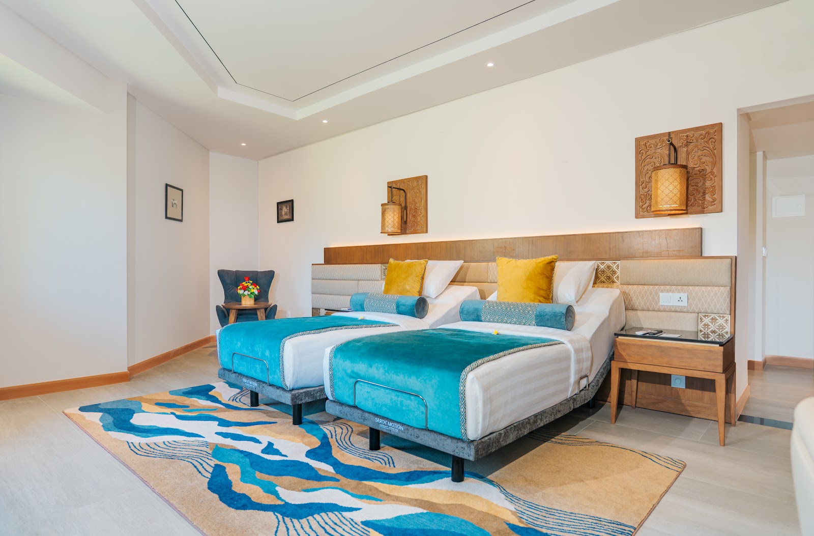 Modern resort suite with two beds topped with turquoise bedding and yellow pillows, wooden headboard, geometric blue and tan rug, and carved wood wall art.
