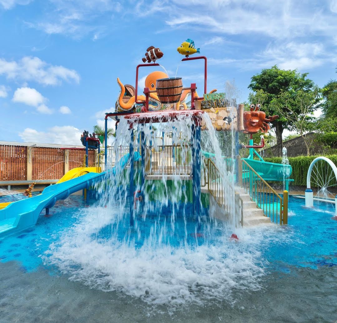 Water cascades from a colorful bucket structure in the Grand Mirage Resort's kids water park, surrounded by slides and tropical trees.