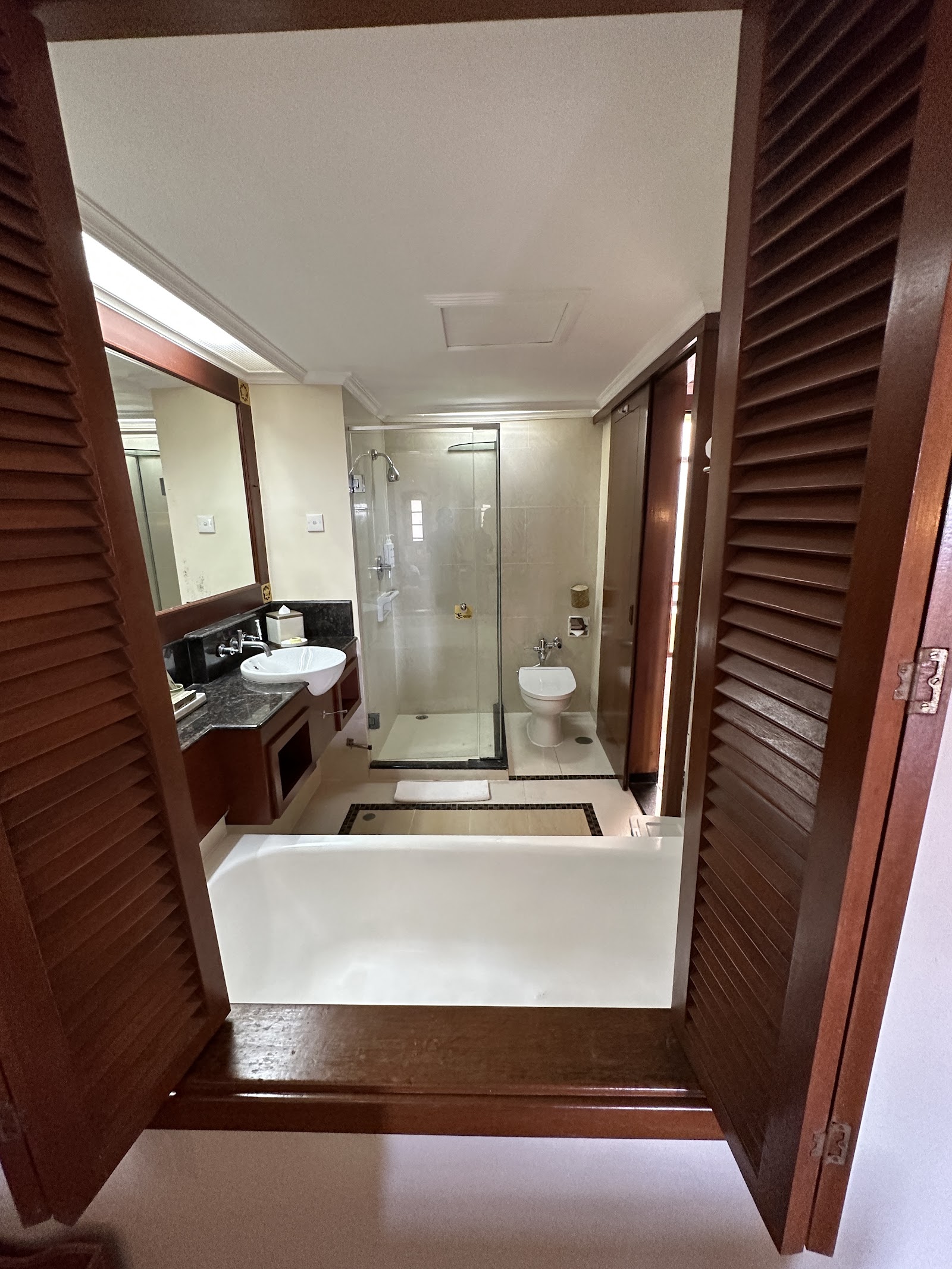 Bathroom with dark wood louvered doors, white soaking tub, walk-in shower with glass enclosure, dual sink vanity with mirror, and toilet.