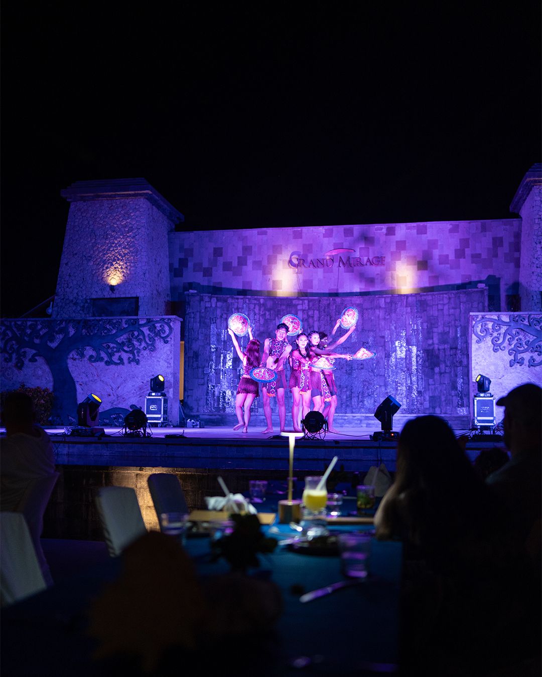 Performers in colorful costumes hold glowing orbs on stage at Grand Mirage Resort during an evening show, with audience members dining in the foreground.
