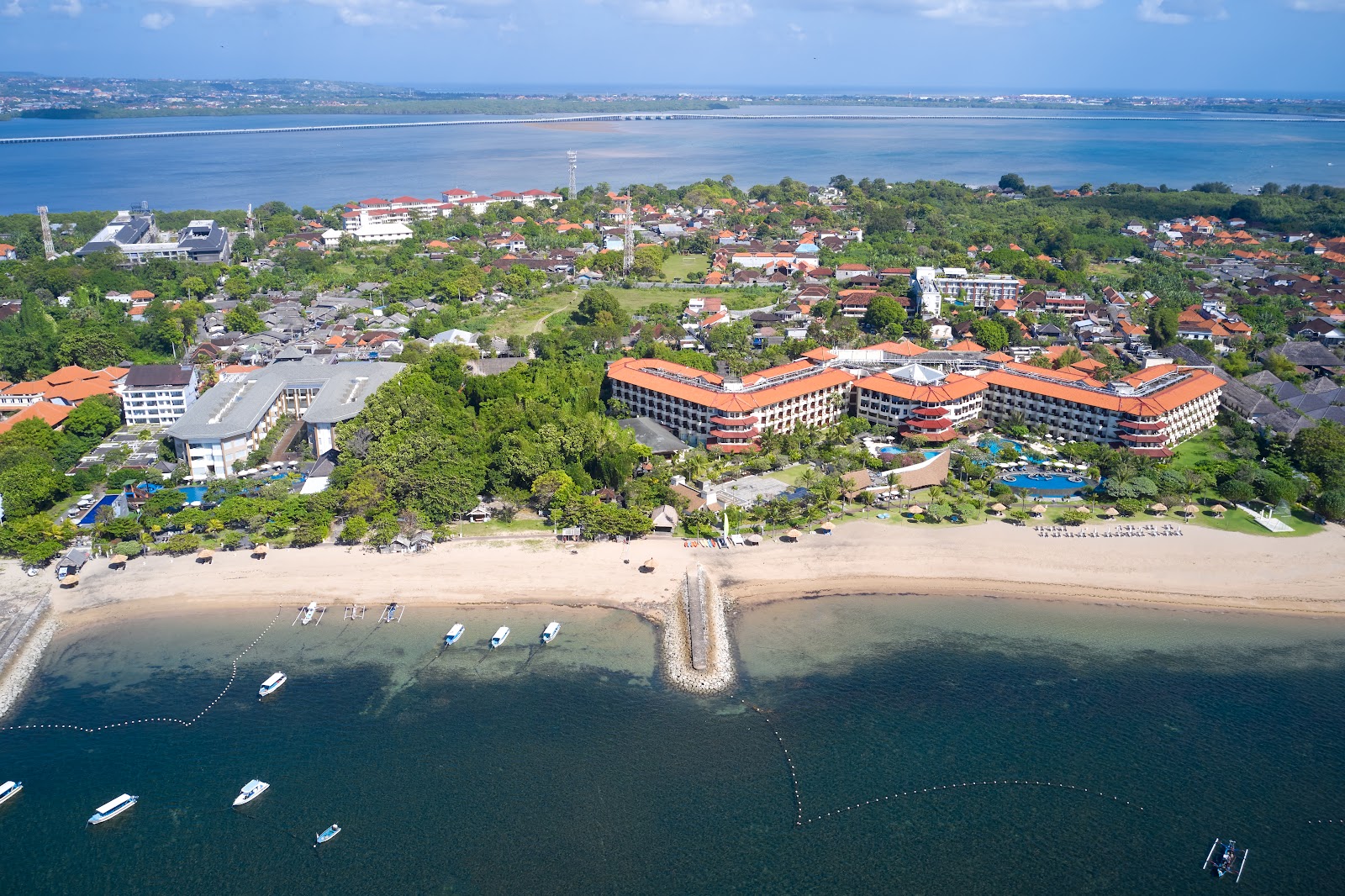 Aerial view of Grand Mirage Resort & Thalasso Bali with orange-roofed buildings, beach, and boats in shallow coastal waters.