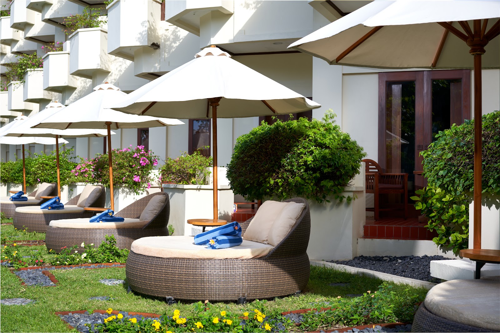 Woven lounge chairs with cushions sit under white umbrellas in the manicured garden courtyard of Grand Mirage Resort & Thalasso Bali, with flowering plants and topiary surrounding the seating area.