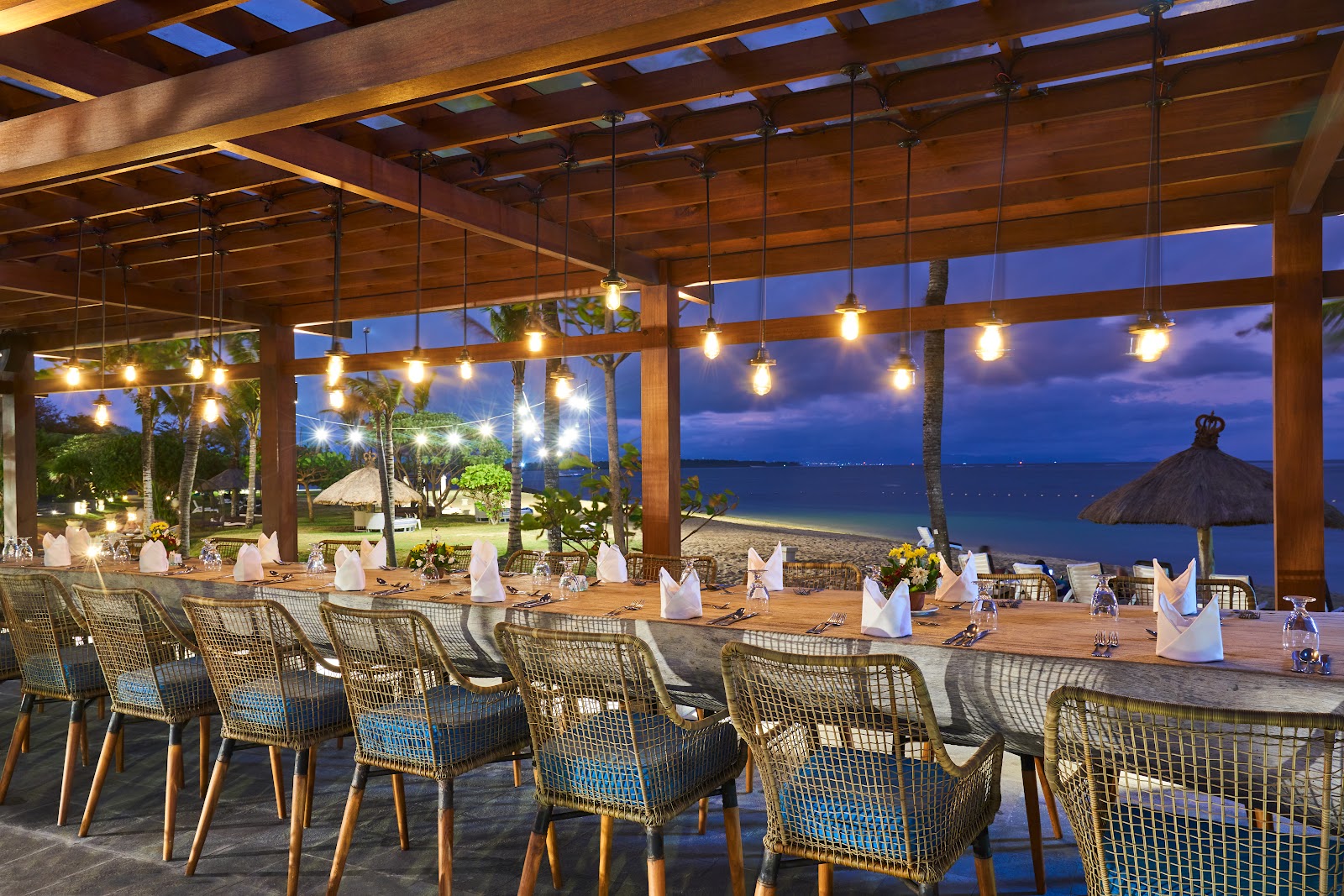 Beachfront dining pavilion at Grand Mirage Resort with woven chairs, long wooden tables set for dinner, and ocean views at dusk.