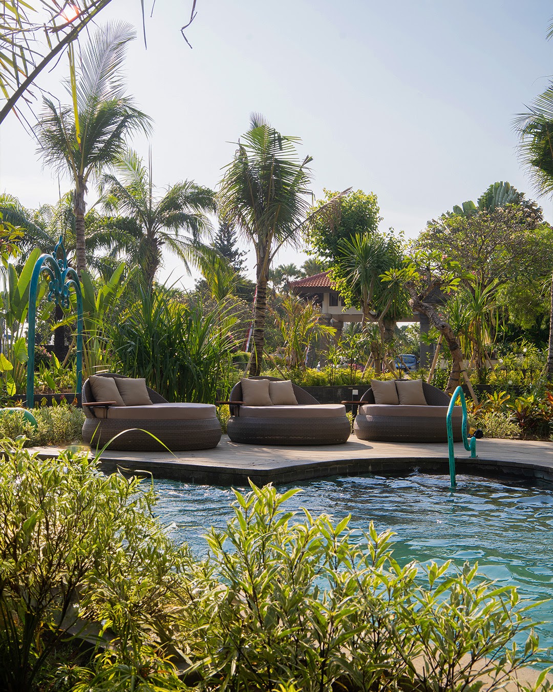 Three round lounge beds on a wooden deck beside a pool, surrounded by lush tropical plants and palm trees at Grand Mirage Resort & Thalasso Bali.