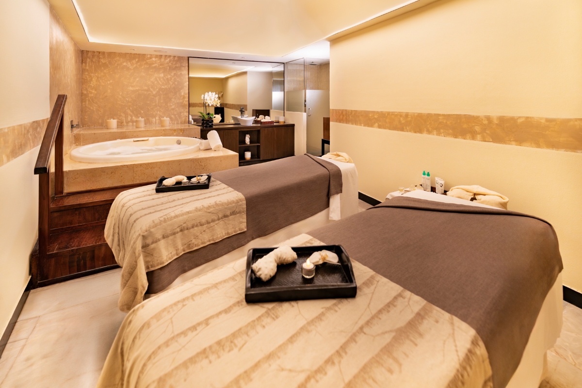Spa treatment room with two massage tables, sunken soaking tub, and dark wood vanity with mirror at Grand Oasis Cancún.