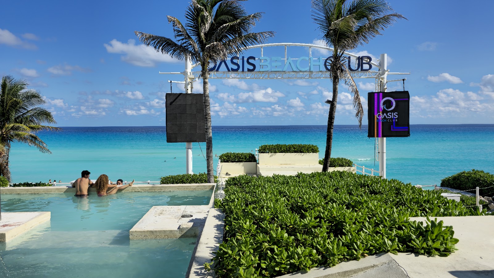 Guests swim in a shallow lagoon at Oasis Beach Club with turquoise ocean and palm trees under blue sky.