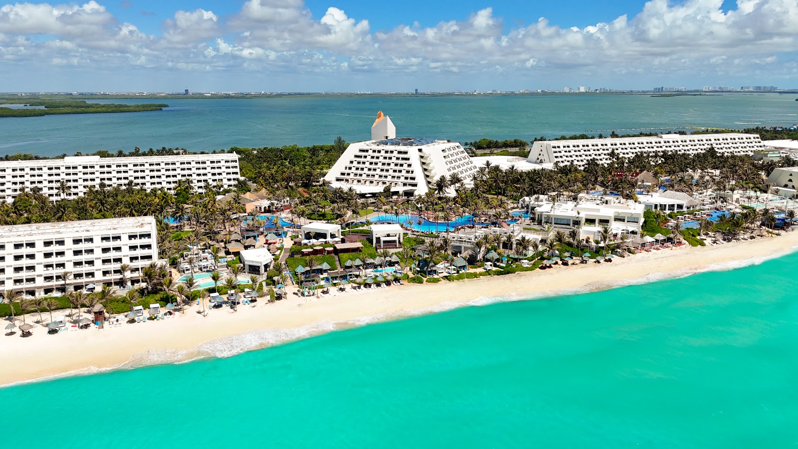 Grand Oasis Cancún sprawls along a turquoise beach with white buildings, pools, and palm trees backed by calm lagoon waters.