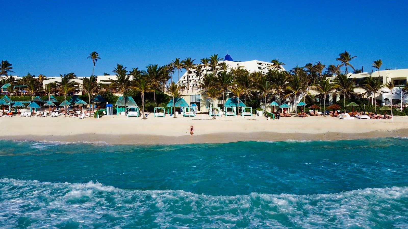 White resort buildings with a blue-domed tower sit behind a sandy beach lined with turquoise umbrellas and palm trees, overlooking the turquoise ocean with rolling waves.