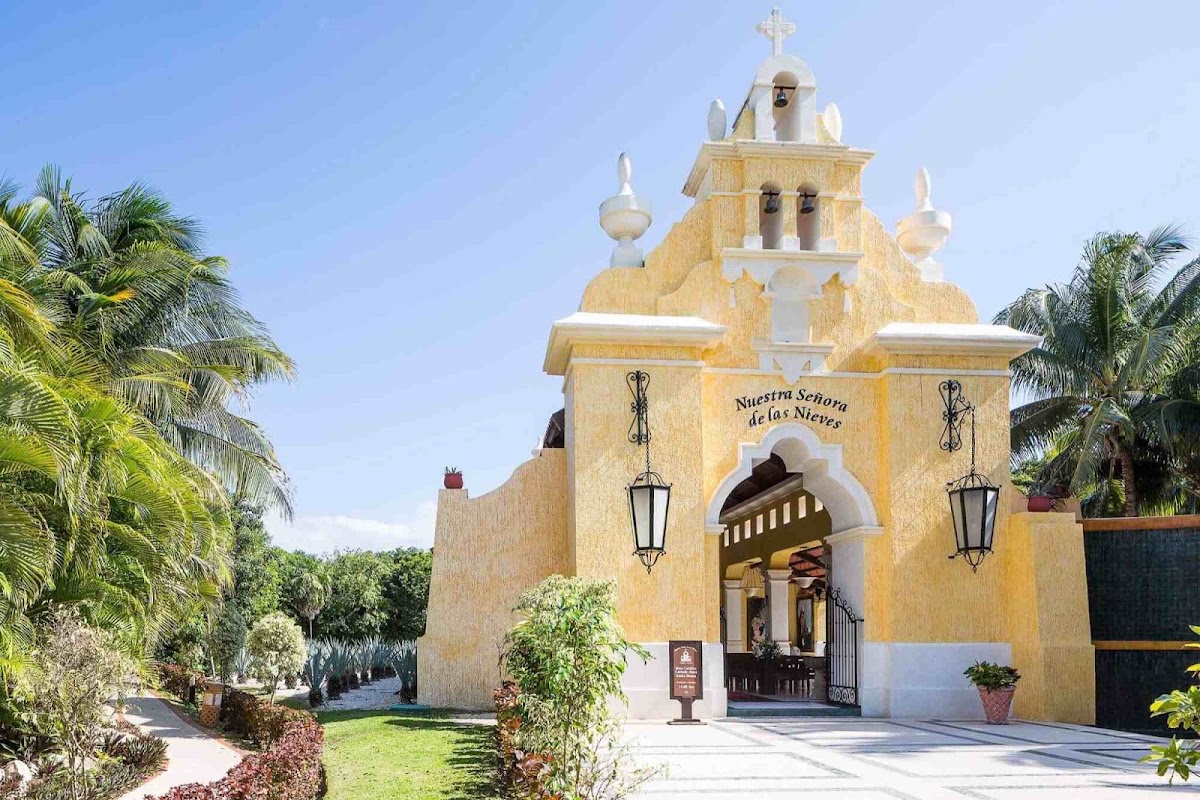 Yellow colonial-style chapel with cross on dome, flanked by palm trees and manicured grounds under clear blue sky.