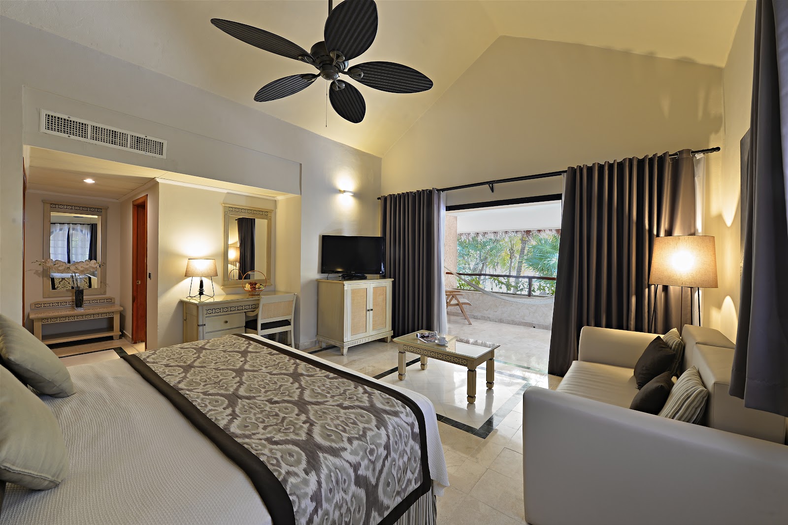 Resort room with bed, sitting area, ceiling fan, and balcony access overlooking tropical grounds.
