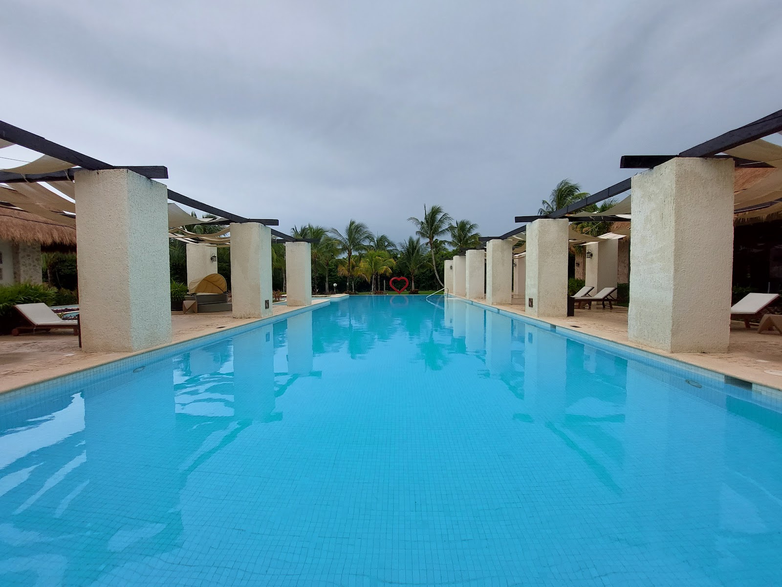 Woman in black swimsuit stands by a curved pool while a guest swims in foreground, with thatched-roof bungalows and white buildings surrounding the Grand Palladium Colonial Resort pool area.