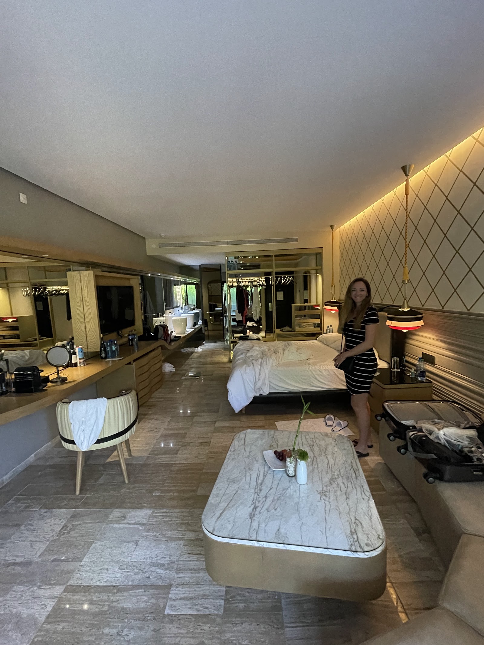 Woman stands in spacious resort suite with marble countertops, dual vanities, beds, and warm ambient lighting at Grand Palladium Colonial Resort & Spa.