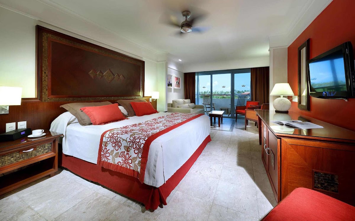Resort suite with king bed, red and brown décor, flat-screen TV, and balcony overlooking tropical grounds.