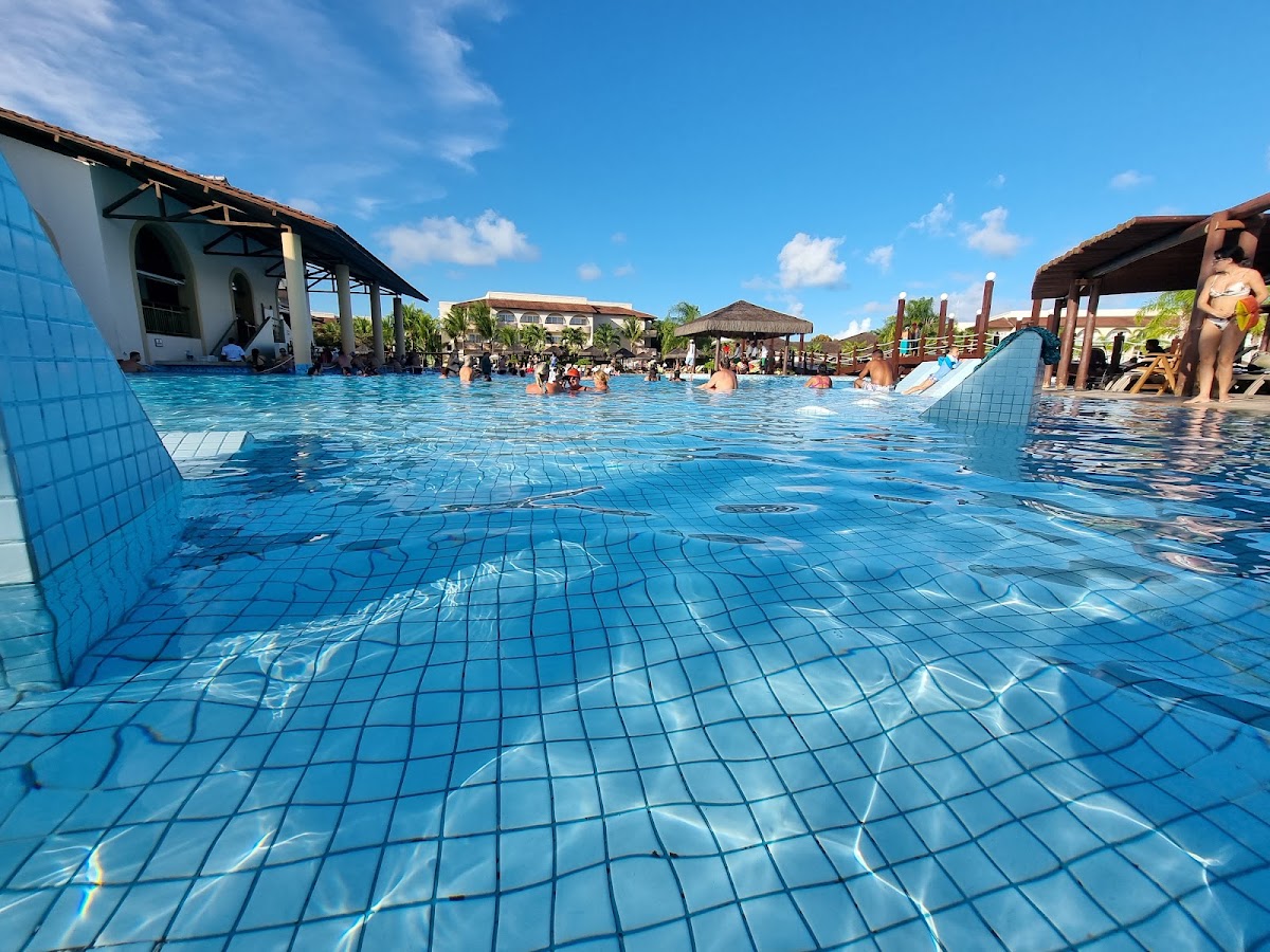 Tiled swimming pool with guests swimming and relaxing, surrounded by thatched-roof structures and palm trees at Grand Palladium Imbassaí.